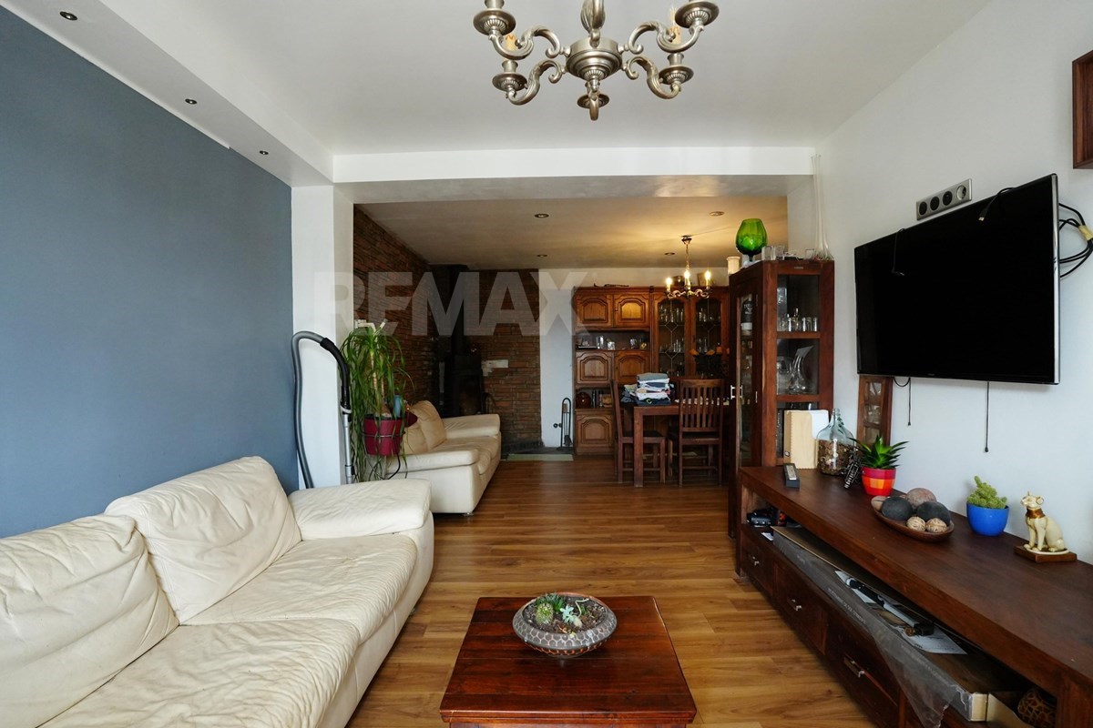 Long living room with cream sofas, a wall-mounted television, wooden media cabinet, coffee table, plants, and an adjoining dining area.