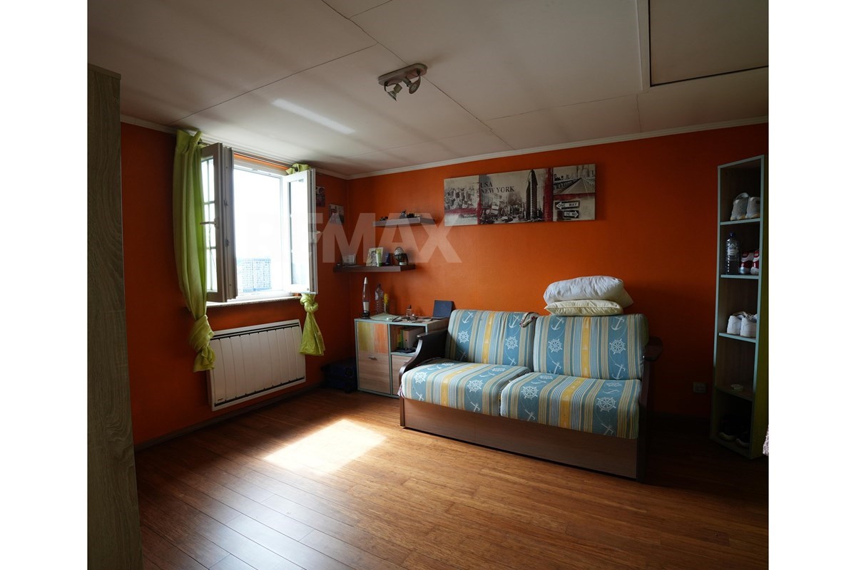 Smaller bedroom with orange walls, striped daybed, open window, radiator, shelves, desk, and a New York-themed wall collage.