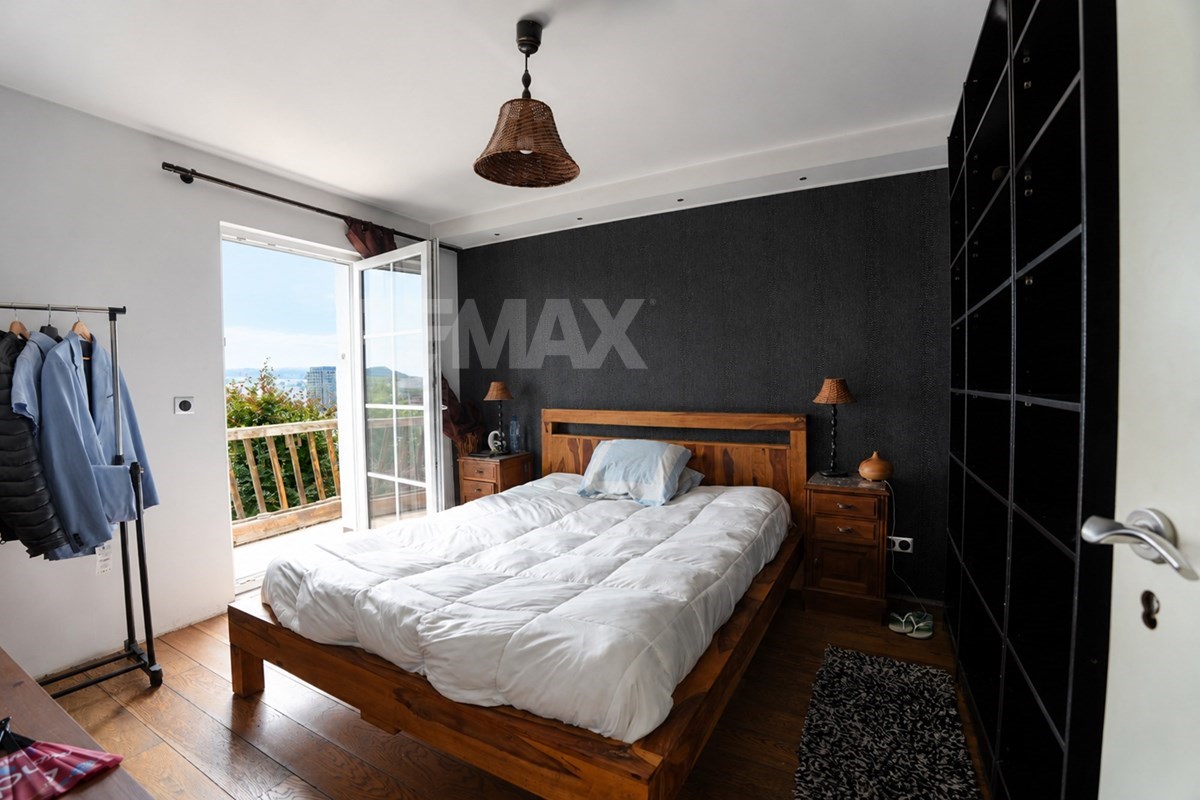 Bedroom with double bed, dark accent wall, bedside cabinets, large black wardrobe, clothing rack, and open balcony doors overlooking distant buildings and hills.
