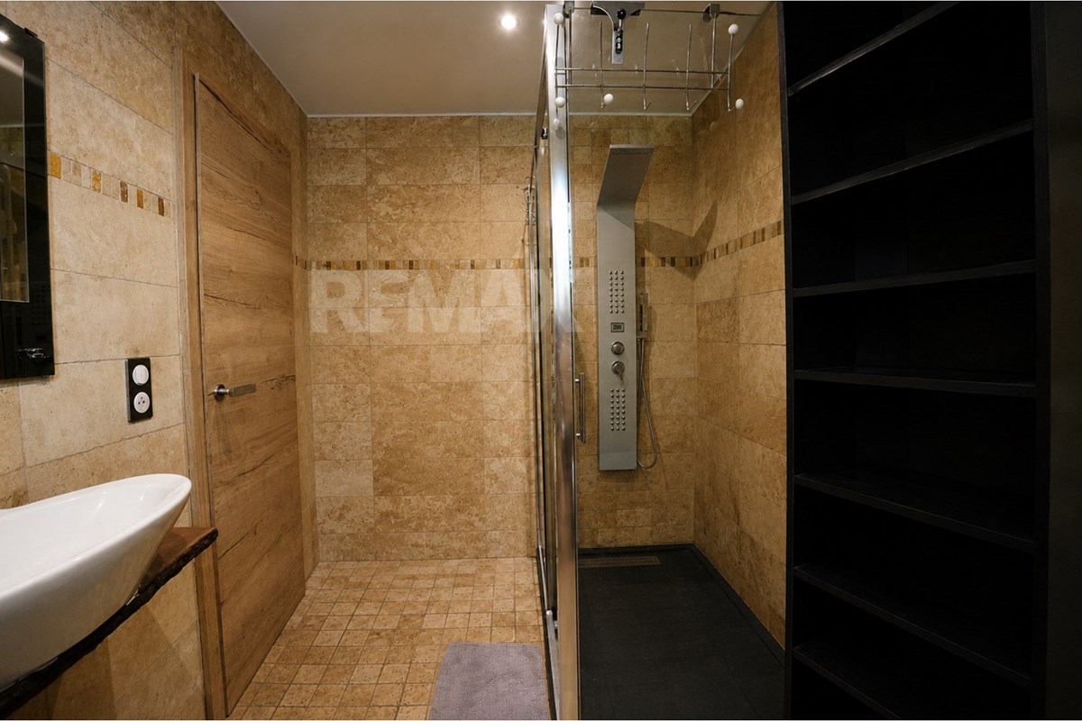 Beige-tiled bathroom with a white basin, glass shower enclosure, rain and hand showers, wooden door, and dark built-in shelving.