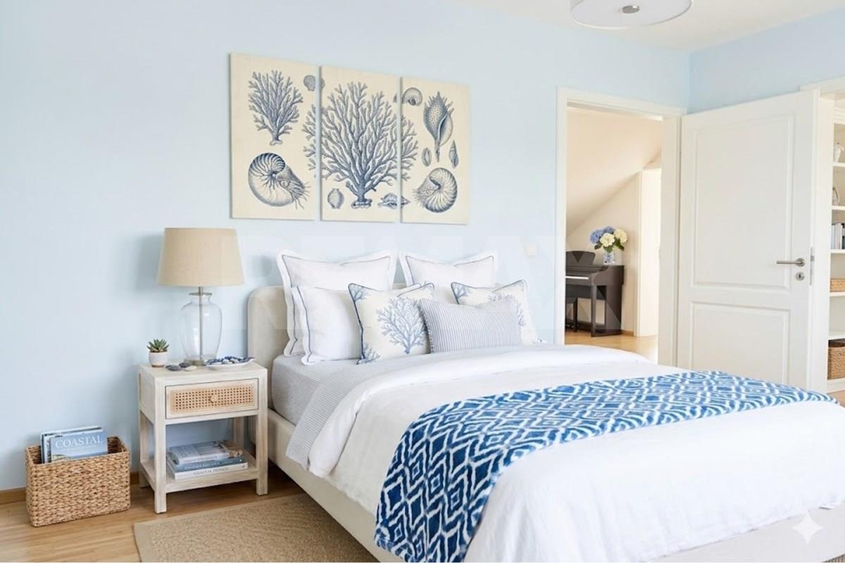 Light blue bedroom with a double bed, coastal shell artwork, bedside lamp, woven storage basket, and an open doorway toward another furnished room.