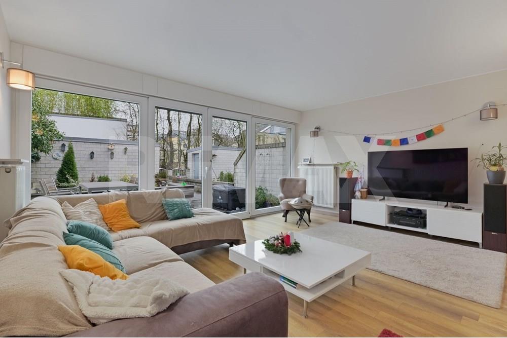 Bright living room with sofa seating, coffee table, television, dining area, and extensive glazing onto a garden.