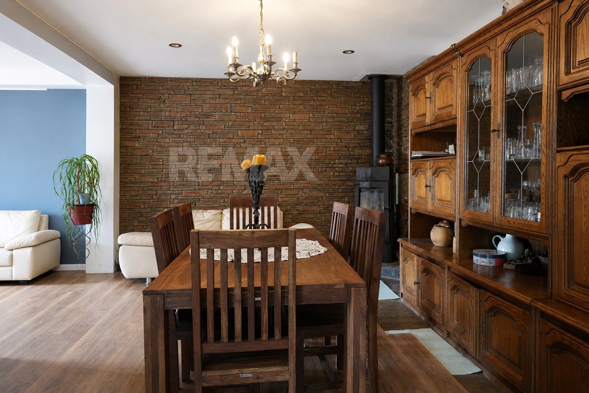 Open dining area with a large wooden table, chairs, chandelier, brick accent wall, wood stove, and glass-fronted wooden cabinet.