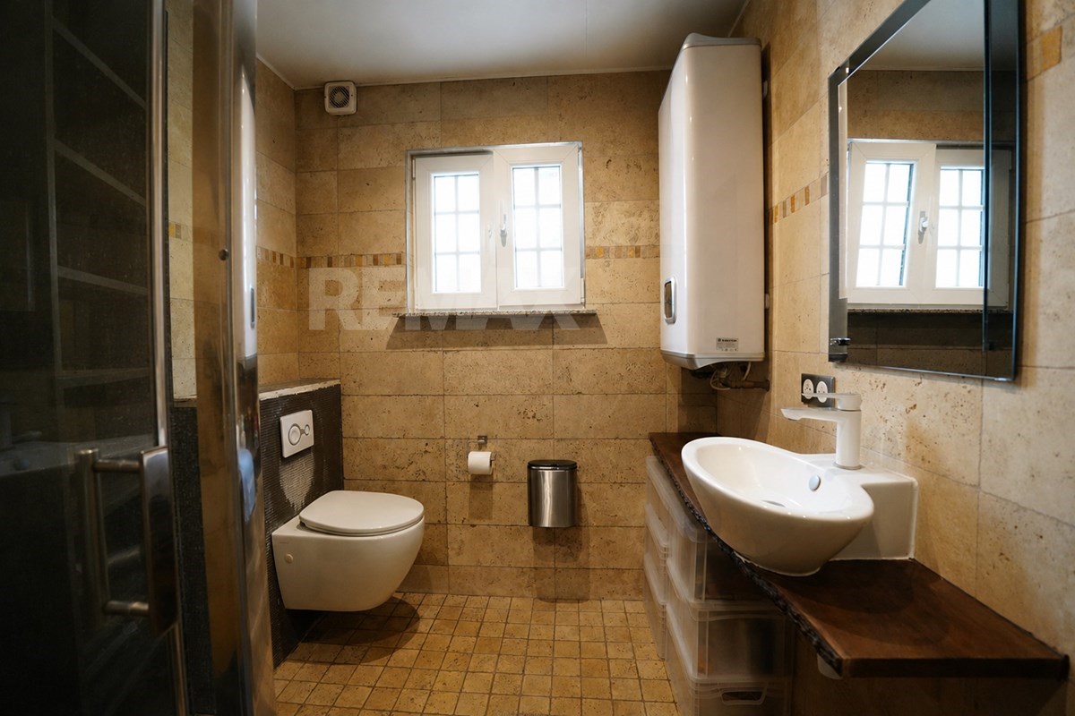 Bathroom with wall-hung toilet, oval vessel basin, large mirror, beige tiles, window radiator, tall water heater, and glass shower edge.